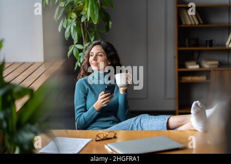 Donna responsabile dei social media lavora da casa seduto al computer portatile chiuso, piedi sulla scrivania bere caffè mattina Foto Stock