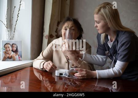 Giovane medico di donna molto abile che mette sulla maschera nebulizzatrice sopra faccia della sua paziente femminile anziana per fare l'inalazione quando visitarlo a casa Foto Stock