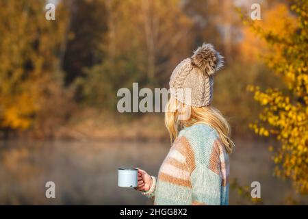 Donna che si rilassa con una bevanda calda accanto al lago in autunno fredda mattina. Campeggio ed escursioni all'aperto nella foresta. Donna capelli biondi con cappello a maglia Foto Stock