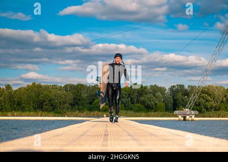 Uomo atletico in muta che porta wakeboard, camminando sul molo dopo l'allenamento Foto Stock