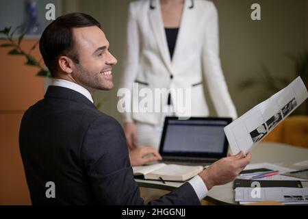 Uomo d'affari in abito classico e camicia bianca si siede alla sua scrivania in Profilo alla fotocamera. Il responsabile dell'azienda delega le attività nel suo ufficio. Colletto bianco Worker .in background è una donna d'affari in un vestito formale bianco. Sfondo burred. Primo piano. Foto di alta qualità Foto Stock