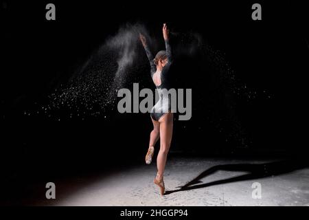 Ballerina flessibile ballerina donna ballare e cospargere la farina su sfondo nero, attraente sottile donna sottile in bodysuit nero in slow motion, gra in movimento Foto Stock