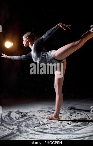 Bella ballerina donna in bodysuit nero balletto da ballo in studio scuro con farina su sfondo nero. Atleta donna che si muove con grazia, Foto Stock