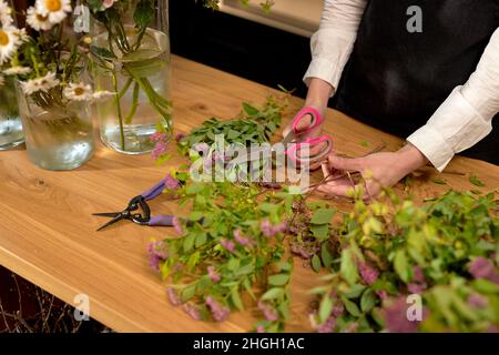 donna che tiene le forbici mentre tagliano i fiori, preparando il bouquet in negozio di fiori. donna irriconoscibile in grembiule nero sta disponendo i petali di fiori selvatici, Foto Stock