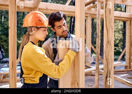 Giovani imprenditori lavoratori di carpentieri in zona di costruzione. Tema industriale. Edificio in legno con struttura a scheletro. Giovane maschio e femmina in Unifor di lavoro Foto Stock