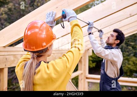 Riparazione di tetti in legno all'aperto. Due falegnami in abiti speciali lavorano in casa nuova. Appaltatori di coperture maschili e femminili che misurano la distanza BE Foto Stock