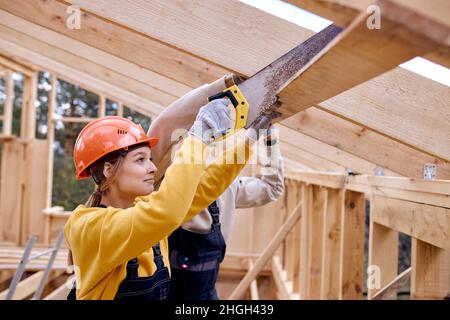 caucasica fiduciosa donna appaltatore in casco arancione con sega taglio a mano trave in legno in casa sito in legno. Giovane uomo reattivo costruttore aiuto e. Foto Stock