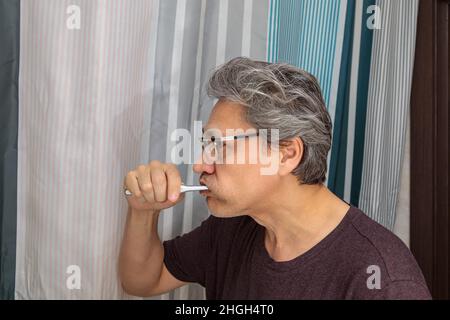 L'uomo adulto dai capelli grigi gli spazzolerà i denti con uno spazzolino in bagno. Foto Stock