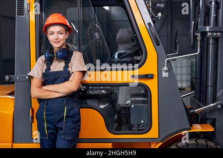 Attraente donna in piedi accanto al conducente del carrello elevatore in un'area industriale in fabbrica, guardando la telecamera con sicurezza, vestito in uniforme. Buon lavoro Foto Stock