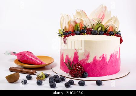 Torta con glassa bianca, mirtilli, frutta su sfondo bianco con sacchetto di pasticceria e spatola, primo piano. Foto Stock