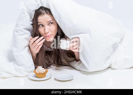 Buongiorno. Camera bianca. Una ragazza carina mangia segretamente la colazione. Una donna si trova a letto con una tazza di bevanda calda, un cupcake è in piedi nelle vicinanze. Foto Stock