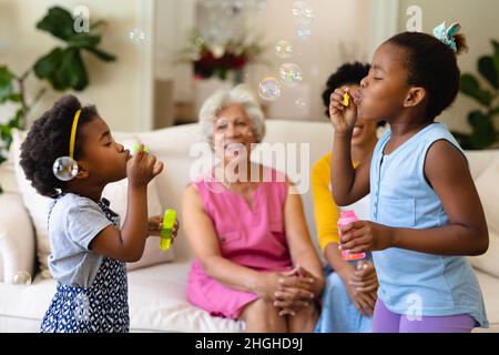 Due sorelle afroamericane che soffiano bolle mentre nonna e madre guardano seduti sul divano Foto Stock