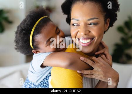 Primo piano della ragazza afroamericana che abbraccia la madre da dietro a casa Foto Stock
