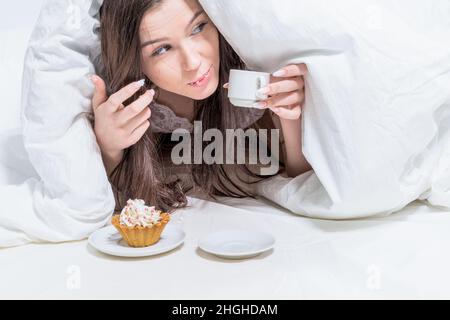 Buongiorno. Camera bianca. La ragazza mangia segretamente la colazione. Una donna è sdraiata a letto, tenendo una tazza di bevanda calda, un cupcake è in piedi nelle vicinanze. Foto Stock