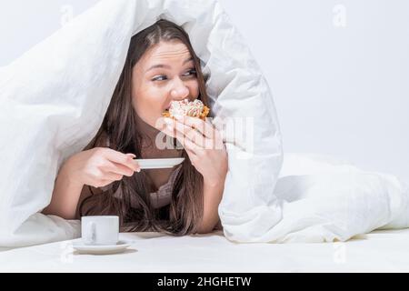 La ragazza mangia segretamente la colazione. Buongiorno. Camera bianca. Una donna è sdraiata a letto mordendo un appetitoso cupcake, c'è un caffè accanto ad esso. Foto Stock