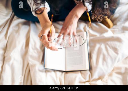 Donna giovane irriconoscibile che mette sui suoi vetri per leggere un libro a letto Foto Stock