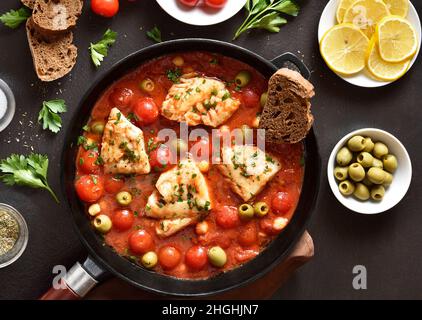 Stufato di merluzzo con ceci, pomodori ciliegini e olive in padella su sfondo di pietra scura. Vista dall'alto, piatto Foto Stock