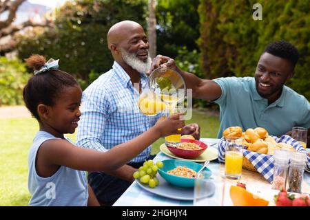 Uomo afroamericano sorridente che serve succo per la figlia da uomo anziano al tavolo da pranzo nel cortile Foto Stock