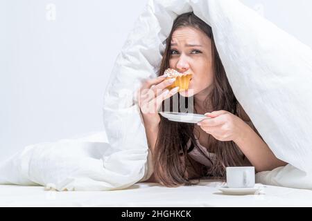 Una donna è sdraiata a letto mordendo un appetitoso cupcake, c'è un caffè accanto ad esso. Buongiorno. Camera bianca. La ragazza mangia segretamente la colazione. Foto Stock