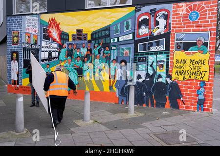 Due uomini trasportano forniture da un furgone bianco e passato un murale che ricorda le rivolte di Brixton 1981 e il razzismo sistematico e le molestie da parte della polizia della comunità nera di Brixton, il 21st gennaio 2022, a Railton Road, Londra, Inghilterra. L'opera, realizzata dagli artisti Jacob V Joyce, Monique Jackson, Ailsa Yexley, Buki Bayode e sola Olulode, è stata coronata da fondi e rappresenta il 40th° anniversario delle rivolte di Brixton del 1981, quando il radicamento è iniziato dal 10th, 11th e 12th aprile 1981 su Brixton's Railton Road, nota come Front Line .. (Didascalia completa in informazioni aggiuntive). Foto Stock