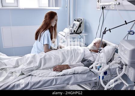 In ospedale Ward recupero Padre è visitato da figlia Adulto. Senior Sick Man dormendo a letto, Redhead figlia caucasica si trova accanto, preoccupante. Foto Stock