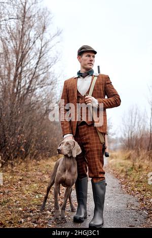 Caucasian Hunter uomo con fucile fucile fucile e con cane da caccia Weimaraner a piedi in strada rurale durante la stagione di caccia. Amichevole cane bello con proprietario in b Foto Stock