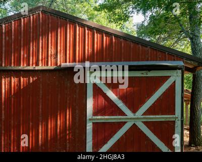 Capannone rosso in azienda ha una porta scorrevole con rivestimento bianco. La luce del sole rivela la presenza di teli di ciottoli polverosi sulla parete. Foto Stock