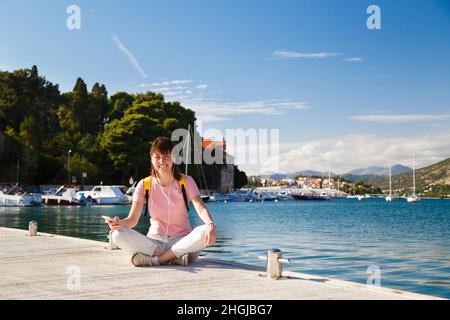 Sorridente giovane femmina turista seduto a gambe incrociate sul molo tenendo cellulare con costa croata in background. Giornata di sole a Dubrovnik, Croazia. Concetto di stile di vita di viaggio Foto Stock