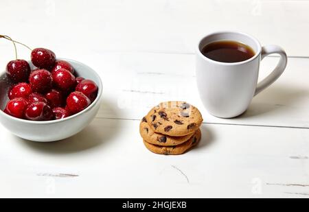 Tazza bianca con caffè, biscotti tradizionali e ciliegie dolci in una ciotola di ceramica su fondo di legno bianco, primo piano. Gustosa colazione, mattina routin Foto Stock