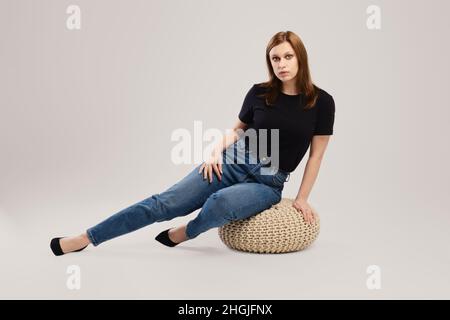 Giovane donna in t shirt casual nera e jeans siede sul pavimento dello studio su sfondo grigio Foto Stock