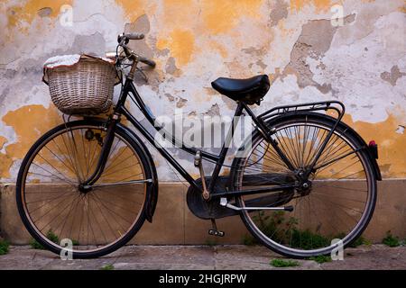 Una vecchia bicicletta con cesto di vimini poggia contro un muro intemperiato a Lucca, Italia Foto Stock