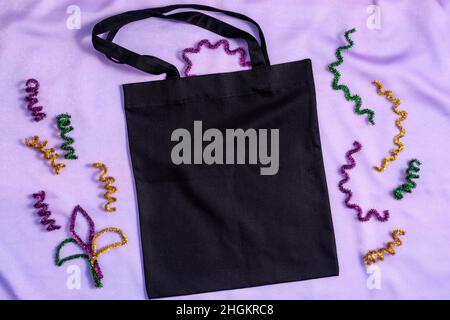 Borsa tote Flatlay Mardi Gras Mockup nera con decori glitterati su sfondo lilla in tessuto. Foto Stock