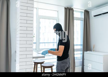 Ladro con balaclava nera rubare laptop. Il burglar commette un crimine in appartamento di lusso Foto Stock