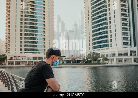 Giovane in maschera medica in piedi all'aperto e guardando avanti verso lo sfondo urbano con skyline della città e grattacieli intorno al lago Foto Stock