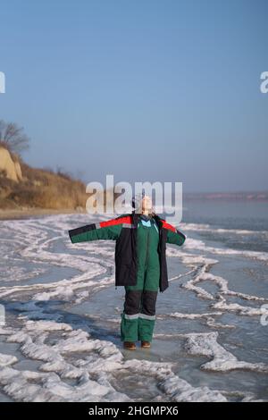 Una donna in un colorato costume termale che gode l'inverno mentre si erge su un lago ghiacciato con cielo blu sullo sfondo. Libertà Foto Stock