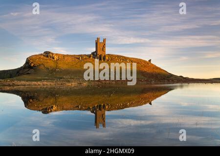 Le rovine della Torre di Lilburn, il Castello di Dunstanburgh sulla costa del Northumberland, si riflettono nelle calme acque di un lago al tramonto in inverno Foto Stock
