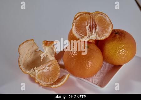 Quattro tangerini sono su una piastra di ceramica bianca e uno è rotto a metà con una pelle parzialmente pelata su uno sfondo bianco. Foto Stock