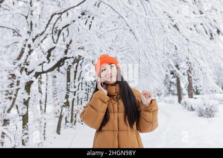 Ritratto di una giovane bella donna asiatica, foto di un primo piano divertirsi parlando al telefono in un parco invernale coperto di neve vestito calorosamente Foto Stock