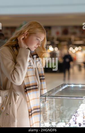 Giovane donna che guarda la vetrina del negozio e gioielli per guardarlo più vicino, bella studentessa modello di moda bionda nel centro commerciale juwellery shopping Foto Stock