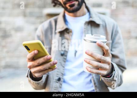 Raccolto anonimo bearded maschio con takeaway bevanda calda in tazza di viaggio. Egli SMS sul cellulare mentre si trova in piedi su strada con shabby edificio durante il viaggio su sfondo sfocato Foto Stock