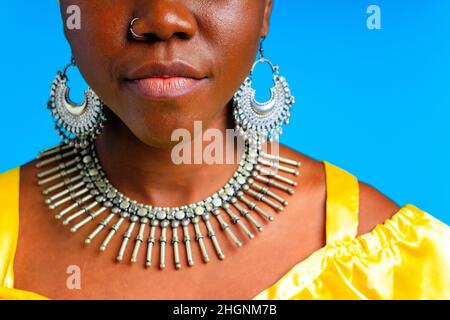 bella donna africana in giallo seta turban sulla testa e vestire con colletto e orecchini argento, naso piercing in blu studio sfondo Foto Stock