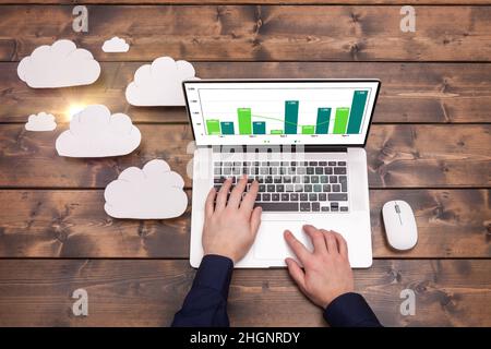 Concetto di tecnologia di cloud computing con nuvole bianche e sole accanto al notebook. Mans mani digitando la tastiera di caricamento dati, su un legno t Foto Stock