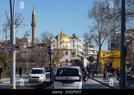 Manavgat, Turchia -20 gennaio 2022: Strada della città con diverse case basse, ponte giallo sul fiume e una moschea, traffico auto attivo, negozi Foto Stock