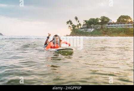 Uomo nero dai capelli lunghi che si paddling su una lunga tavola da surf al punto di surf nell'oceano Indiano. Palmeto ha littato raggi del tramonto sullo sfondo. Acqua estrema s Foto Stock