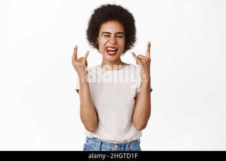 Rock on. Sorridendo ragazza africana con capelli afro, mostrando sassy segno di metallo pesante, rock sul gesto, godendo e divertirsi, in piedi su sfondo bianco Foto Stock