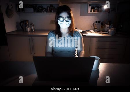 Una ragazza in una maschera e guanti impara durante la quarantena. Una donna che lavora fuori orario da casa su un notebook. Un freelance è seduto in cucina nel Foto Stock