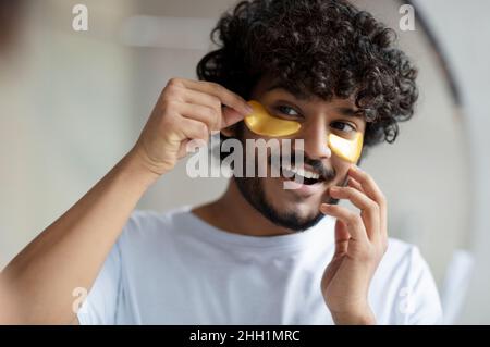 Cura della pelle per ridurre i sacchetti per gli occhi. L'uomo indiano allegro che applica i cerotti anti-affaticamento dell'oro sotto gli occhi, guardando lo specchio Foto Stock