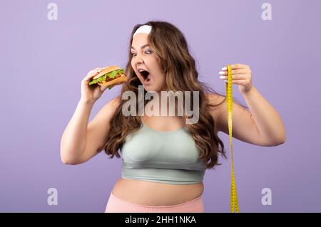 Divertente affamato caucasico più giovane taglia in abbigliamento sportivo con nastro di misurazione mangia appetitoso hamburger Foto Stock