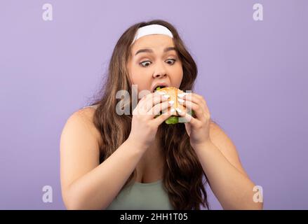 Divertente fame caucasica più dimensione giovane femmina in uniforme sport mangia golosamente hamburger Foto Stock