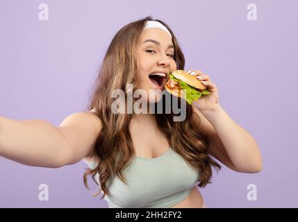 Divertente sorridente abbastanza affamato caucasico più taglia giovane donna in uniforme sport mangiare hamburger e prendere selfie Foto Stock